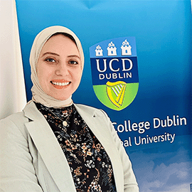 A woman stands in front of the UCD logo and smiles at the camera. She is wearing a cream blazer and floral shirt.